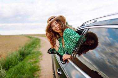 Araba camından sarkan şapkalı ve parlak elbiseli bir gezgin. Şapkalı güzel bir kadın araba yolculuğundan zevk alıyor, özgürlüğü hissediyor. Seyahat ve eğlence konsepti. Etkin yaşam biçimi.