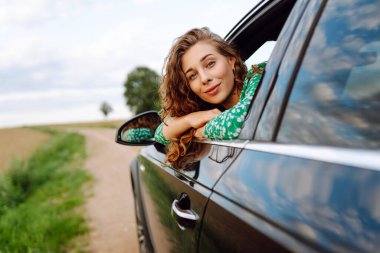 Araba camından sarkan şapkalı ve parlak elbiseli bir gezgin. Şapkalı güzel bir kadın araba yolculuğundan zevk alıyor, özgürlüğü hissediyor. Seyahat ve eğlence konsepti. Etkin yaşam biçimi.