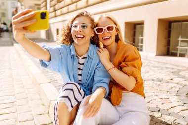İki neşeli kadın güneşli caddelerde telefonlarıyla selfie çekiyor. Güzel bayan turistler birlikte dışarıda vakit geçiriyorlar. Arkadaşlık, eğlence ve turizm konsepti.