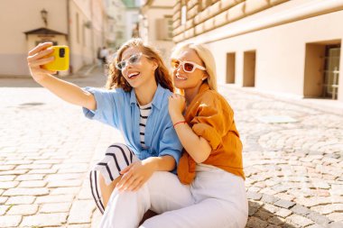 İki neşeli kadın güneşli caddelerde telefonlarıyla selfie çekiyor. Güzel bayan turistler birlikte dışarıda vakit geçiriyorlar. Arkadaşlık, eğlence ve turizm konsepti.