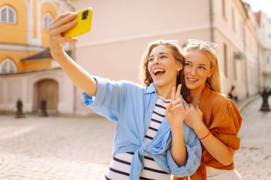 İki neşeli kadın güneşli caddelerde telefonlarıyla selfie çekiyor. Güzel bayan turistler birlikte dışarıda vakit geçiriyorlar. Arkadaşlık, eğlence ve turizm konsepti.
