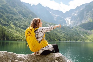 Renkli bir sırt çantasıyla bir kadın yürüyüşçü dağ gölü manzarasından zevk alır ve özgürlüğü hisseder. Genç bir kadın dağlarda yürüyüş yolunda, bir taşın üzerinde oturuyor. Yürüyüş ve doğa konsepti.