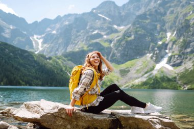 Renkli bir sırt çantasıyla bir kadın yürüyüşçü dağ gölü manzarasından zevk alır ve özgürlüğü hisseder. Genç bir kadın dağlarda yürüyüş yolunda, bir taşın üzerinde oturuyor. Yürüyüş ve doğa konsepti.