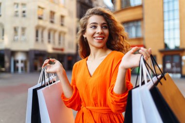 Güneşli caddede duran alışveriş çantalı hoş bir kadın. Alışverişten sonra yürüyüş yapmaktan hoşlanan genç bir kadın. Tüketim ve yürüme kavramı.