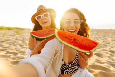 Mutlu bayan arkadaşlar gün batımı kumsalında karpuz yiyip eğleniyorlar. İki güzel kadın birlikte vakit geçirir ve deniz kıyısında piknik yaparlar. Eğlence ve arkadaşlık kavramı.