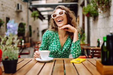 Samimi verandadaki ahşap masada kahve içen mutlu kadının portresi. Genç bir turist sabah keyfine bakıyor ve sıcak bir şeyler içiyor. Turizm ve içki konsepti.
