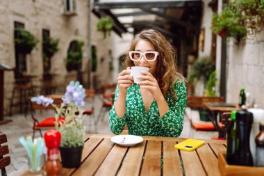 Samimi verandadaki ahşap masada kahve içen mutlu kadının portresi. Genç bir turist sabah keyfine bakıyor ve sıcak bir şeyler içiyor. Turizm ve içki konsepti.