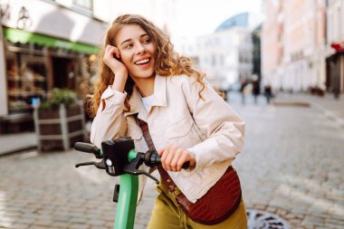 Güneşli şehir sokaklarında elektrikli scooter kullanan neşeli bir kadın. Güzel bir kadın dinleniyor ve dışarı çıkmaktan zevk alıyor. Ekoloji ve turizm konsepti. Etkin yaşam biçimi.