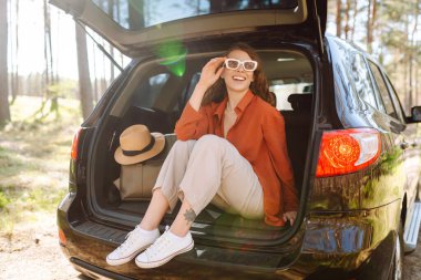 Genç bir kadın arabanın bagajında oturur ve güneşli bir ormanda havanın tadını çıkarır. Bir araba macerasında gezgin arabada zevk ve rahatlık hisseder. Doğa konsepti ve yolculuk.