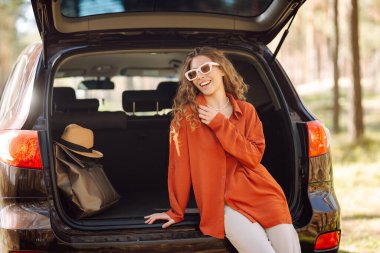 Genç bir kadın arabanın bagajında oturur ve güneşli bir ormanda havanın tadını çıkarır. Bir araba macerasında gezgin arabada zevk ve rahatlık hisseder. Doğa konsepti ve yolculuk.