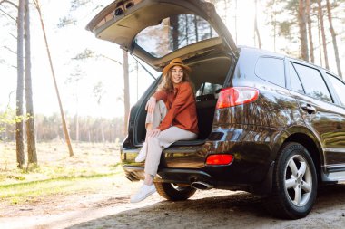 Genç bir kadın arabanın bagajında oturur ve güneşli bir ormanda havanın tadını çıkarır. Bir araba macerasında gezgin arabada zevk ve rahatlık hisseder. Doğa konsepti ve yolculuk.