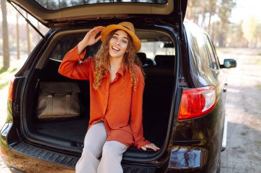 Genç bir kadın arabanın bagajında oturur ve güneşli bir ormanda havanın tadını çıkarır. Bir araba macerasında gezgin arabada zevk ve rahatlık hisseder. Doğa konsepti ve yolculuk.