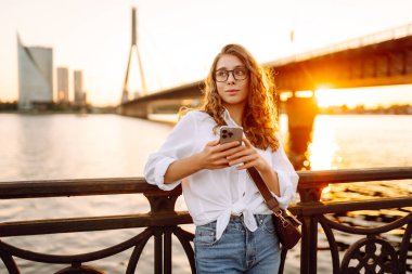 Gün batımında elinde telefon ve gözlüklü kıvırcık kadın sette yürüyor. Akıllı telefonlu genç bir kadın havanın tadını çıkarıyor. Yürüme ve rahatlama konsepti.