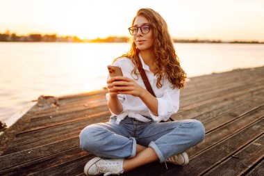 Gün batımında elinde telefon ve gözlüklü kıvırcık kadın sette yürüyor. Akıllı telefonlu genç bir kadın havanın tadını çıkarıyor. Yürüme ve rahatlama konsepti.