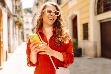 Güneşli şehir sokaklarında telefonla yürüyen genç bir kadının portresi. Güzel bir kadın seyahat etmekten, blog yazmaktan ya da telefonda mesajlaşmaktan hoşlanıyor. Turizm, hafta sonu ve teknoloji konsepti.