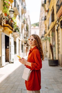 Güneşli şehir sokaklarında telefonla yürüyen genç bir kadının portresi. Güzel bir kadın seyahat etmekten, blog yazmaktan ya da telefonda mesajlaşmaktan hoşlanıyor. Turizm, hafta sonu ve teknoloji konsepti.