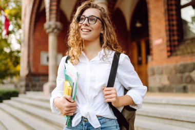 Gözlüklü ve kitaplı güzel bir öğrenci üniversite kampüsü geçmişine karşı duruyor. Gün batımında havanın tadını çıkaran kitap ve defterlerle dolu hoş bir kadın. Eğitim konsepti. Yaşam biçimi.
