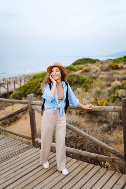 Şapkalı ve sırt çantalı güzel bir kadın ahşap yürüyüş yolunda yürüyor ve deniz manzarasının tadını çıkarıyor. Genç turistler doğadan zevk alıyorlar. Seyahat ve tatil konsepti. Etkin yaşam biçimi.