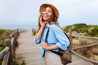 Şapkalı ve sırt çantalı güzel bir kadın ahşap yürüyüş yolunda yürüyor ve deniz manzarasının tadını çıkarıyor. Genç turistler doğadan zevk alıyorlar. Seyahat ve tatil konsepti. Etkin yaşam biçimi.