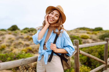 Şapkalı ve sırt çantalı güzel bir kadın ahşap yürüyüş yolunda yürüyor ve deniz manzarasının tadını çıkarıyor. Genç turistler doğadan zevk alıyorlar. Seyahat ve tatil konsepti. Etkin yaşam biçimi.