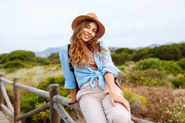 Şapkalı ve sırt çantalı güzel bir kadın ahşap yürüyüş yolunda yürüyor ve deniz manzarasının tadını çıkarıyor. Genç turistler doğadan zevk alıyorlar. Seyahat ve tatil konsepti. Etkin yaşam biçimi.