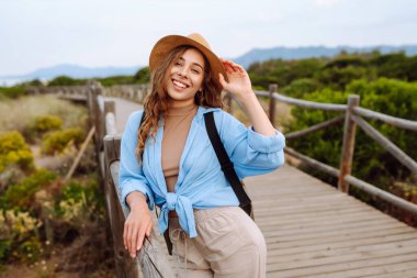 Şapkalı ve sırt çantalı güzel bir kadın ahşap yürüyüş yolunda yürüyor ve deniz manzarasının tadını çıkarıyor. Genç turistler doğadan zevk alıyorlar. Seyahat ve tatil konsepti. Etkin yaşam biçimi.