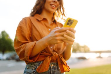 Gün batımında elinde telefon tutan genç bir kadının yakın çekimi. Dışarıda telefonla konuşan gülümseyen bir kadın. Teknoloji kavramı, blog yazma. Yaşam biçimi.