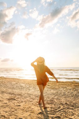 Gün batımında sahilde yürüyen genç bir kadın. Arkasında deniz manzarası olan, özgür hisseden ve günbatımının tadını çıkaran güzel bir kadın. Doğa, rahatlama ve keyif konsepti.