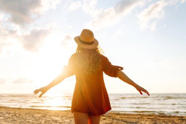 Gün batımında sahilde yürüyen genç bir kadın. Arkasında deniz manzarası olan, özgür hisseden ve günbatımının tadını çıkaran güzel bir kadın. Doğa, rahatlama ve keyif konsepti.