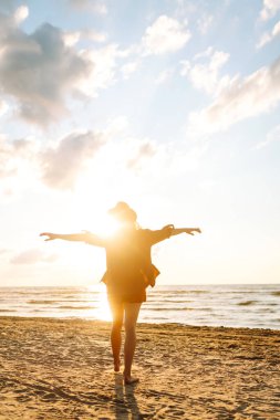 Gün batımında sahilde yürüyen genç bir kadın. Arkasında deniz manzarası olan, özgür hisseden ve günbatımının tadını çıkaran güzel bir kadın. Doğa, rahatlama ve keyif konsepti.