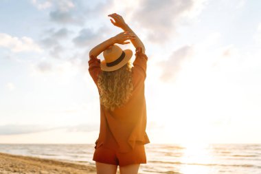 Gün batımında sahilde yürüyen genç bir kadın. Arkasında deniz manzarası olan, özgür hisseden ve günbatımının tadını çıkaran güzel bir kadın. Doğa, rahatlama ve keyif konsepti.