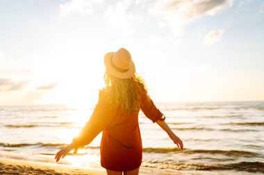 Gün batımında sahilde yürüyen genç bir kadın. Arkasında deniz manzarası olan, özgür hisseden ve günbatımının tadını çıkaran güzel bir kadın. Doğa, rahatlama ve keyif konsepti.