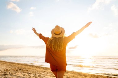 Gün batımında sahilde yürüyen genç bir kadın. Arkasında deniz manzarası olan, özgür hisseden ve günbatımının tadını çıkaran güzel bir kadın. Doğa, rahatlama ve keyif konsepti.