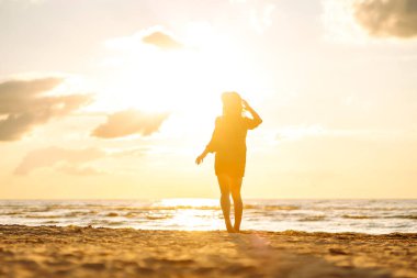 Gün batımında sahilde yürüyen genç bir kadın. Arkasında deniz manzarası olan, özgür hisseden ve günbatımının tadını çıkaran güzel bir kadın. Doğa, rahatlama ve keyif konsepti.