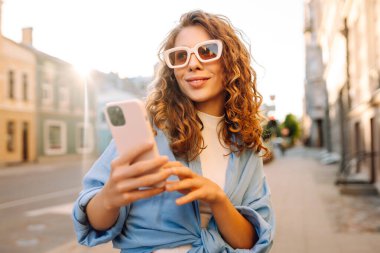 Güneş gözlüklü ve telefonlu mutlu kadın güneşli şehir sokaklarında yürüyor. Dışarıda blog yazan ve mesajlaşan genç bir kadın. Etkin yaşam biçimi.