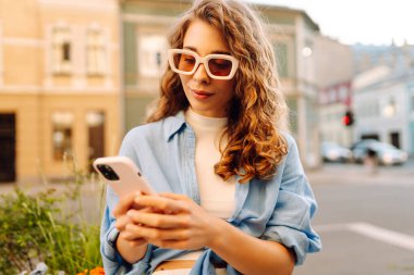 Güneş gözlüklü ve telefonlu mutlu kadın güneşli şehir sokaklarında yürüyor. Dışarıda blog yazan ve mesajlaşan genç bir kadın. Etkin yaşam biçimi.