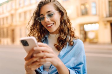 Güneş gözlüklü ve telefonlu mutlu kadın güneşli şehir sokaklarında yürüyor. Dışarıda blog yazan ve mesajlaşan genç bir kadın. Etkin yaşam biçimi.