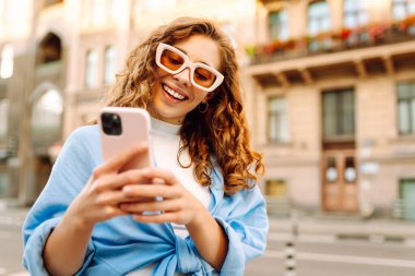Güneş gözlüklü ve telefonlu mutlu kadın güneşli şehir sokaklarında yürüyor. Dışarıda blog yazan ve mesajlaşan genç bir kadın. Etkin yaşam biçimi.