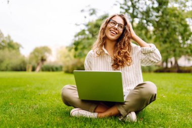 Yeşil çimlerin üzerinde dizüstü bilgisayarı güneş ışınlarıyla oturan neşeli kadın. Güzel bayan serbest yazar dizüstü bilgisayarda çalışmaktan ve dışarıda dinlenmekten zevk alıyor. Uzak çalışma konsepti.