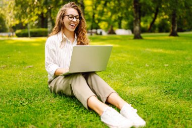 Yeşil çimlerin üzerinde dizüstü bilgisayarı güneş ışınlarıyla oturan neşeli kadın. Güzel bayan serbest yazar dizüstü bilgisayarda çalışmaktan ve dışarıda dinlenmekten zevk alıyor. Uzak çalışma konsepti.