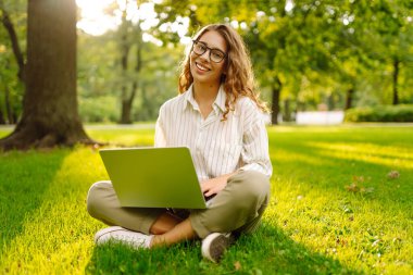 Yeşil çimlerin üzerinde dizüstü bilgisayarı güneş ışınlarıyla oturan neşeli kadın. Güzel bayan serbest yazar dizüstü bilgisayarda çalışmaktan ve dışarıda dinlenmekten zevk alıyor. Uzak çalışma konsepti.