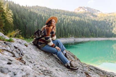 Şapka takan ve dağ gölünün tadını çıkaran sırt çantalı kadın gezginin arka görüntüsü. Yürüyüşün tadını çıkaran, güneşli bir günde dağlarda özgürlüğünü hisseden genç bir kadın. Seyahat, doğa konsepti..