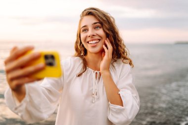 Elinde telefon olan genç bir kadının portresi arka planda deniz olan selfie çekiyor. Güzel kadın blogları ya da günbatımının tadını çıkarırken telefonuyla görüntülü konferanslar. Boş zaman ve günlük konsepti.