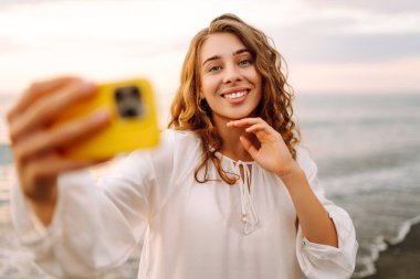 Elinde telefon olan genç bir kadının portresi arka planda deniz olan selfie çekiyor. Güzel kadın blogları ya da günbatımının tadını çıkarırken telefonuyla görüntülü konferanslar. Boş zaman ve günlük konsepti.