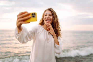 Elinde telefon olan genç bir kadının portresi arka planda deniz olan selfie çekiyor. Güzel kadın blogları ya da günbatımının tadını çıkarırken telefonuyla görüntülü konferanslar. Boş zaman ve günlük konsepti.
