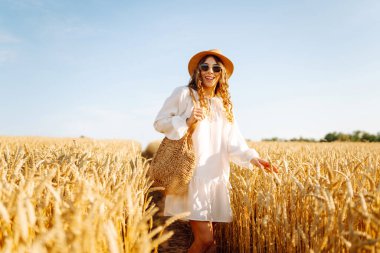 Genç bir kadın altın buğday tarlasında güneşli bir günün tadını çıkarıyor. Beyaz elbiseli ve şapkalı güzel bir kadın tarlada yürüyor, zevk alıyor. Yürümek, doğa konsepti..