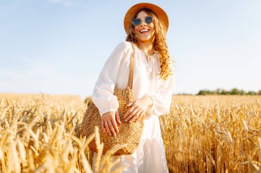 Genç bir kadın altın buğday tarlasında güneşli bir günün tadını çıkarıyor. Beyaz elbiseli ve şapkalı güzel bir kadın tarlada yürüyor, zevk alıyor. Yürümek, doğa konsepti..