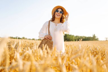 Genç bir kadın altın buğday tarlasında güneşli bir günün tadını çıkarıyor. Beyaz elbiseli ve şapkalı güzel bir kadın tarlada yürüyor, zevk alıyor. Yürümek, doğa konsepti..