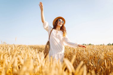 Genç bir kadın altın buğday tarlasında güneşli bir günün tadını çıkarıyor. Beyaz elbiseli ve şapkalı güzel bir kadın tarlada yürüyor, zevk alıyor. Yürümek, doğa konsepti..