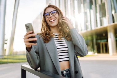 Şık giyinmiş, elinde telefonla modern binanın yakınında güneşli bir günün tadını çıkaran genç bir kadın. Şık bir kadın akıllı telefona bakıyor ve dışarıda gülümsüyor. Keyif ve rahatlama konsepti. Yaşam biçimi.